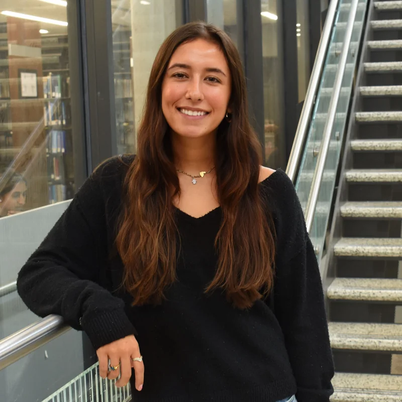 Olivia Catani smiling, with long brown hair and a black sweater, leaning on a glass railing beside a staircase inside a campus building.